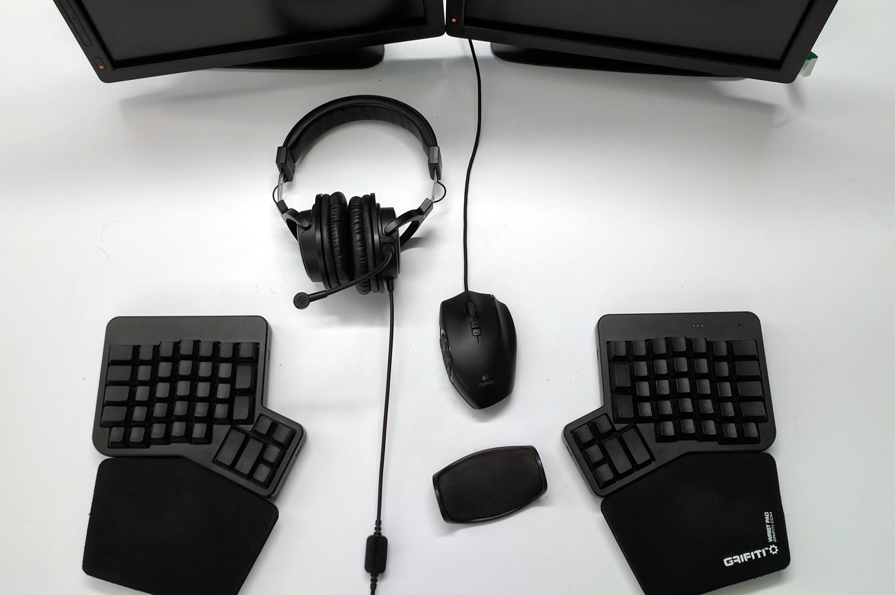 Two halves of a split keyboard flank both sides of a mouse and a pair of headphones against the backdrop of a stark white desk surface.